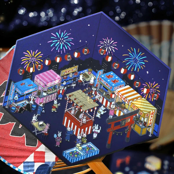 Colorful paper model of a Japanese festival scene with stalls and fireworks.