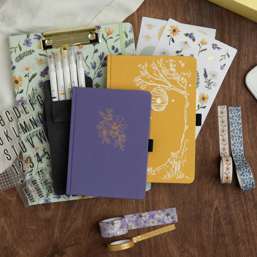 Collection of notebooks, stationery items, and decorative ribbons on a wooden surface. June Quarterly Box Beekeeper’s Garden from Archer & Olive.