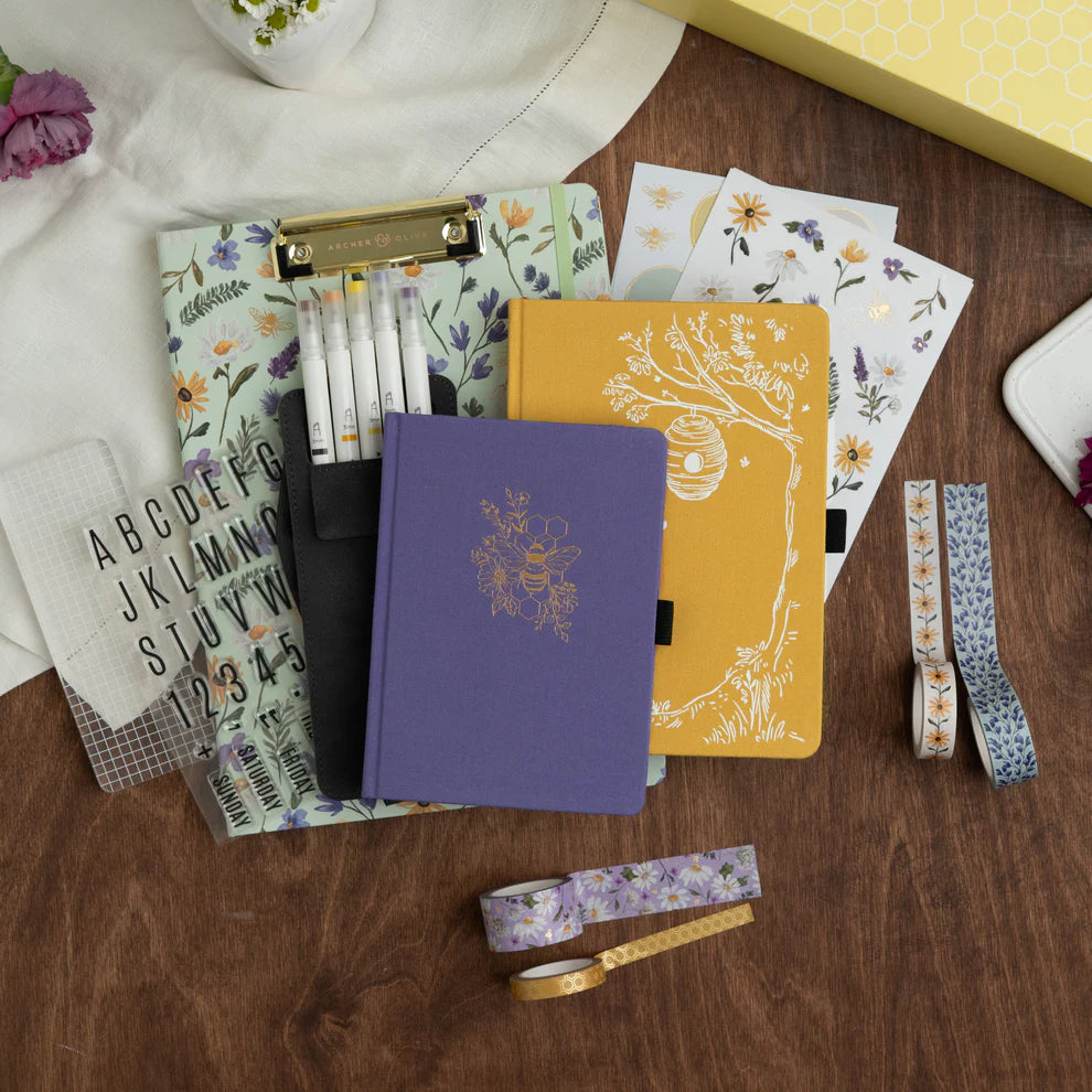 Collection of notebooks, stationery items, and decorative ribbons on a wooden surface. June Quarterly Box Beekeeper’s Garden from Archer & Olive.
