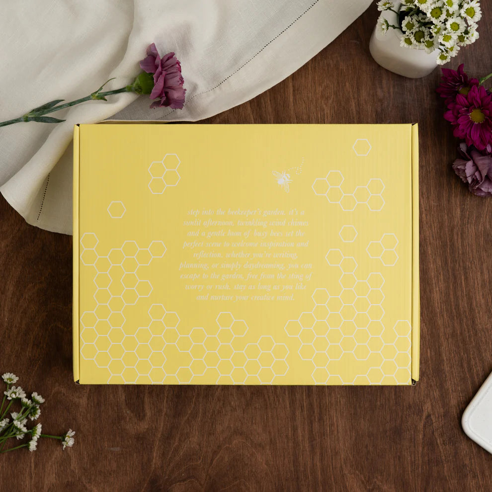 Yellow box with floral patterns on a wooden surface with flowers around. June Quarterly Box Beekeeper’s Garden from Archer & Olive.